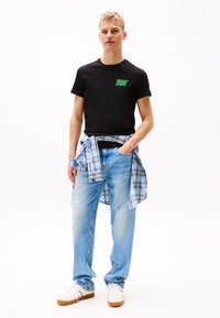 Tricou negru din bumbac cu logo verde, asociat cu blugi denim de un albastru deschis. O cămașă în carouri albastre este legată în jurul taliei. Adidași albi.