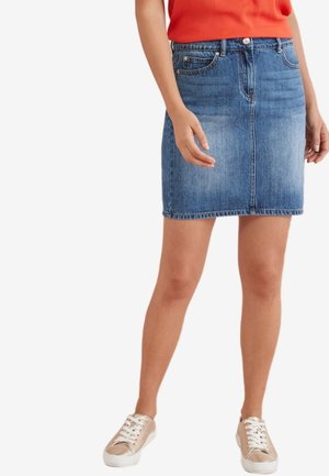 Kvinde iført en blå denimnederdel, orange top og beige sneakers, stående med hænderne afslappet ned langs siderne.