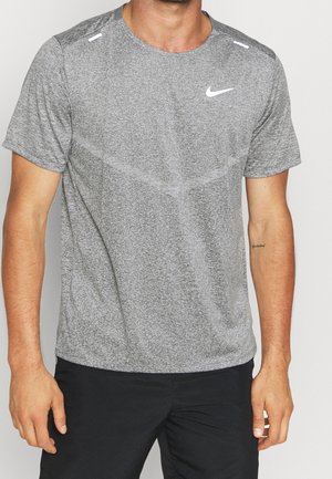 Homme portant un t-shirt de sport Nike gris à manches courtes et un pantalon noir, debout devant un fond clair uni.