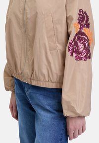 Beige bomberjack met een ritssluiting aan de voorkant, elastische manchetten en bloemrijke pailletteborduursels in roze en oranje op de rechtermouw.