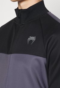 Chaqueta negra y gris con cremallera hasta el cuello, con una textura suave y un logotipo gris en el pecho.