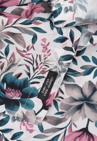 Bloemprint stof met grote teal en roze bloemen met groene bladeren op een witte achtergrond. Inclusief een zwart label met tekst.