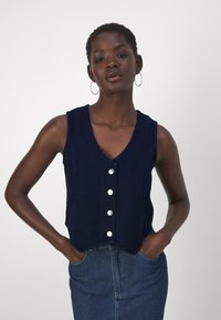 Ναυτικό μπλε αμάνικο τοπ με V-ντεκολτέ, πέντε μεγάλους ασημένιους κουμπιά μπροστά και ελαφρώς καμπυλωτή πορεία. Η υφή φαίνεται λεία.