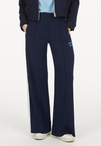 Marinefarbene weite Beinhosen aus weichem Stoff, mit einem blauen Logo auf der Tasche und weißen Seitenstreifen. Elastischer Bund und Seitentaschen.