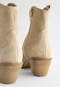 Stivali alla caviglia in suede beige con punta appuntita, dettagli laterali testurizzati e tacco affusolato di 5 cm, dotati di una cerniera dorata sul retro.