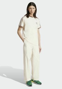 Κρεμ μπλουζάκι με καφέ λεπτομέρειες και τρεις ρίγες, συνδυασμένο με κρεμ παντελόνι. Slim fit, μαλακό ύφασμα, μινιμαλιστικό λογότυπο Adidas στο στήθος.