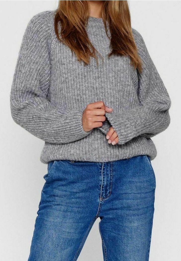 Femme portant un pull en tricot gris et un jean bleu, tenant la manche du pull d'une main près de la taille.
