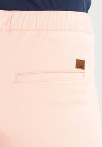 Světlé růžové kalhoty se zadním našitým kapsou a malou hnědou koženou nášivkou se značkou "Roxy" na pásku kalhot.