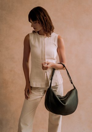 Woman in beige sleeveless top and pants holding a large black leather handbag against a plain beige background.