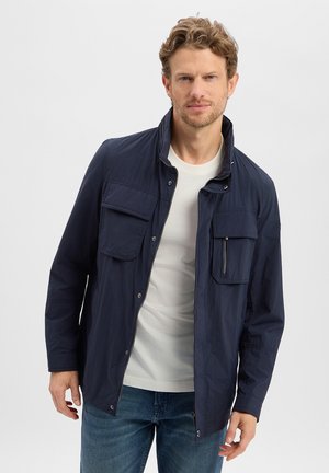 Mężczyzna z krótkimi kręconymi włosami, ubrany w granatową kurtkę na białą koszulę i niebieskie dżinsy, stoi na tle jednolitego jasnego tła.