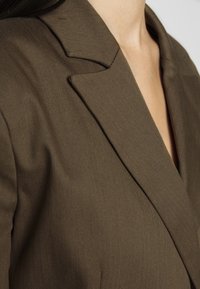 Hnědý blazer s výstřihem do V, strukturovaným designem a texturovanou látkou, který ukazuje přiléhavý fit s hlubokým výstřihem.