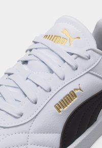 Λευκό αθλητικό παπούτσι Puma με χρυσό λογότυπο Puma στη γλώσσα και στη πλευρά, μαύρες λεπτομέρειες και λευκά κορδόνια δεμένα σε σχέδιο σταυρωτό.