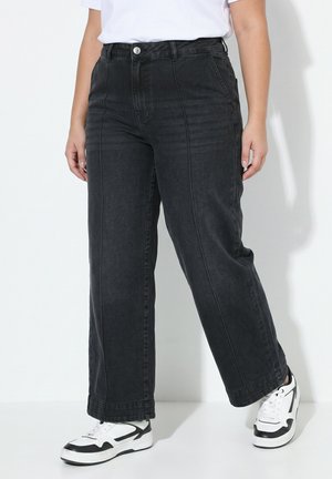 Person iført sorte vidde jeans og hvide sneakers med sorte detaljer, stående mod en ensfarvet hvid baggrund.