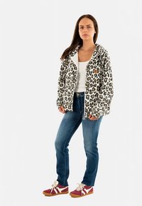 Giacca con zip e cappuccio con stampa leopardata, base bianca e macchie marroni. Indossata sopra una maglietta bianca e abbinata a jeans blu e scarpe da ginnastica rosse.