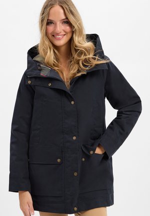 Lachende blonde vrouw met een zwarte capuchonjas met drukknopen en steekzakken, staande tegen een effen witte achtergrond.