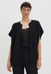 Haut noir ample avec un col rond, des manches courtes et une ouverture à l'avant. Porté par-dessus un haut noir transparent et associé à un pantalon noir.