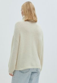 Cremfarbener, gestrickter Pullover mit lockerer Passform und hohem Kragen. Weiche Textur, lange Ärmel und abgerundetem Saum. Über hellblauen Jeans getragen.