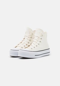 Wysokie trampki z płótna w kolorze ecru z białymi gumowymi podeszwami i czarnymi paskami, wyposażone w okrągłe sznurówki i metalowe przelotki.