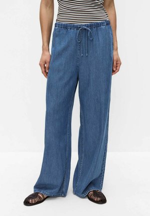 Personne portant un pantalon en denim bleu à jambes larges avec une taille à cordon, des sandales tressées noires et un haut rayé noir et blanc.