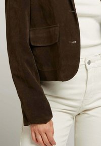 Gros plan sur une personne portant une veste en suede marron avec une poche à rabat et un pantalon blanc taille haute, la main reposant sur le côté.