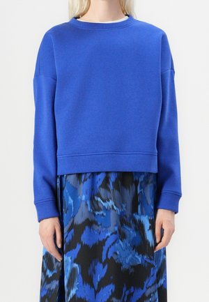 Person iført en knaldblå langærmet sweatshirt kombineret med et blåt og sort nederdel med abstrakt mønster.