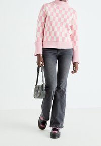 Suéter de cuello alto a rayas rosa y crema con puños acanalados, combinado con jeans de campana negros desgastados y zapatos de plataforma negros, sosteniendo un bolso gris.