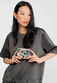 Cartera con cremallera multicolor que presenta estampados abstractos en azul, dorado y negro sobre un fondo beige, sostenida contra una parte superior gris carbón.