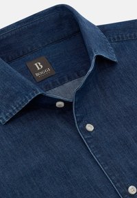 Ciemnoniebieska koszula jeansowa z klasycznym kołnierzykiem, białymi guzikami i czarną metką z nazwą marki "BOGGI MILANO". Gładka tekstura.