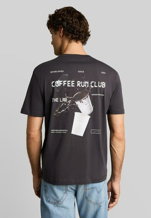 Man draagt een zwart T-shirt met een afbeelding van twee koffiekopjes die omvallen en tekst "Coffee Run Club" op de rug, gecombineerd met lichtblauwe spijkerbroek.