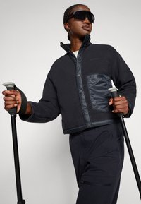 Persoon in een zwarte jas en donkere broek, die wandelstokken vasthoudt, omhoog kijkt, grote zwarte zonnebril draagt, tegen een effen achtergrond.