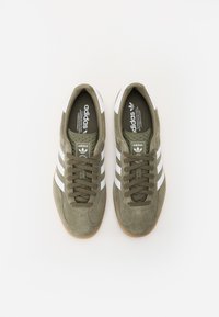 Adidași din suede verde măsliniu cu dungi albe, design cu șireturi, suprafață texturată și talpă din cauciuc natural. Branding pe limbă și pe toc.
