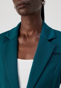 Närbild av en mörkhyad person som bär en teal kavaj över en vit ribbad topp, med silverfärgade ringörhängen och flätat hår som är uppsatt.