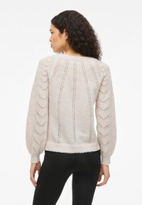 Γυναίκα με πλάτη στραμμένη, φορώντας ένα κρεμ πλεκτό πουλόβερ με ανοιχτές κάθετες ρίγες και φουσκωτά μανίκια, συνδυασμένο με μαύρο παντελόνι.