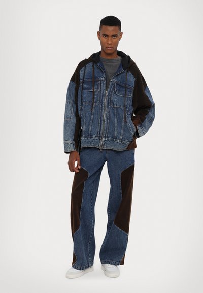 Denimjakke med hætte, der har en blanding af blå nuancer og mørkebrune lapper. Kombinationen af brede denimbukser og hvide sneakers.