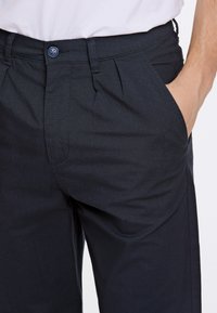 Pantaloni blu navy in tessuto testurizzato, con pieghe, una tasca laterale e un bottone blu in vita per la chiusura.