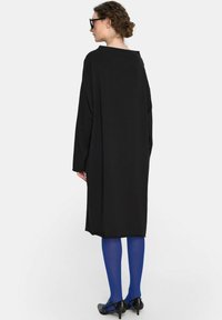 Czarna długa sukienka o luźnym kroju, szerokimi rękawami i okrągłym dekoltem. Połączona z niebieskimi rajstopami i czarnymi butami na obcasie.