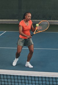 Tennis player in an orange short-sleeve shirt and gray shorts, holding an orange racket with a yellow ball, on a blue court.