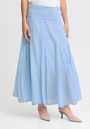 Personne portant une jupe évasée bleu clair, longue jusqu’aux chevilles avec une large ceinture, et des escarpins slingback beiges à talons, debout devant un fond uni.