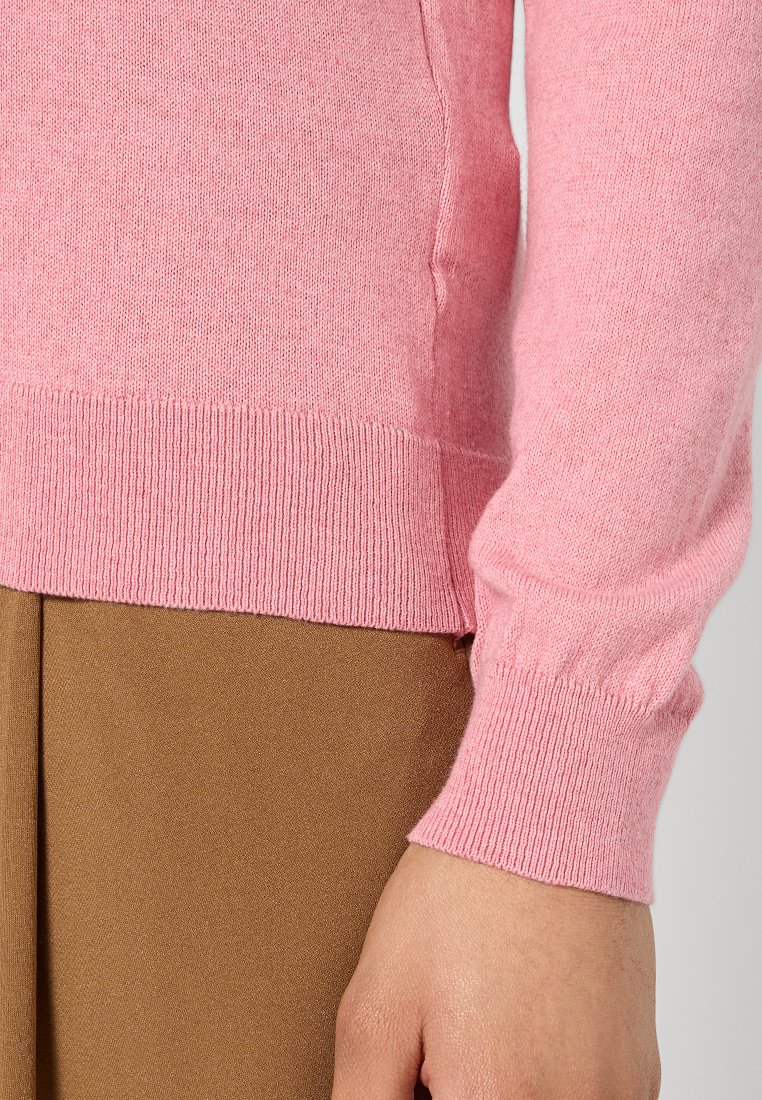 Maglione a maglia rosa chiaro con polsini e fondo a coste, abbinato a un pantalone in tessuto marrone. La morbidezza del tessuto e il design aderente sono evidenti.