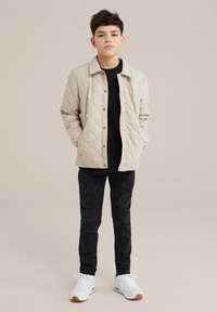 Beige pumpattu takki, jossa on nepparit, päällä musta paita. Mustat slim-fit-housut ja valkoiset tossut ruskeilla yksityiskohdilla täydentävät asun.