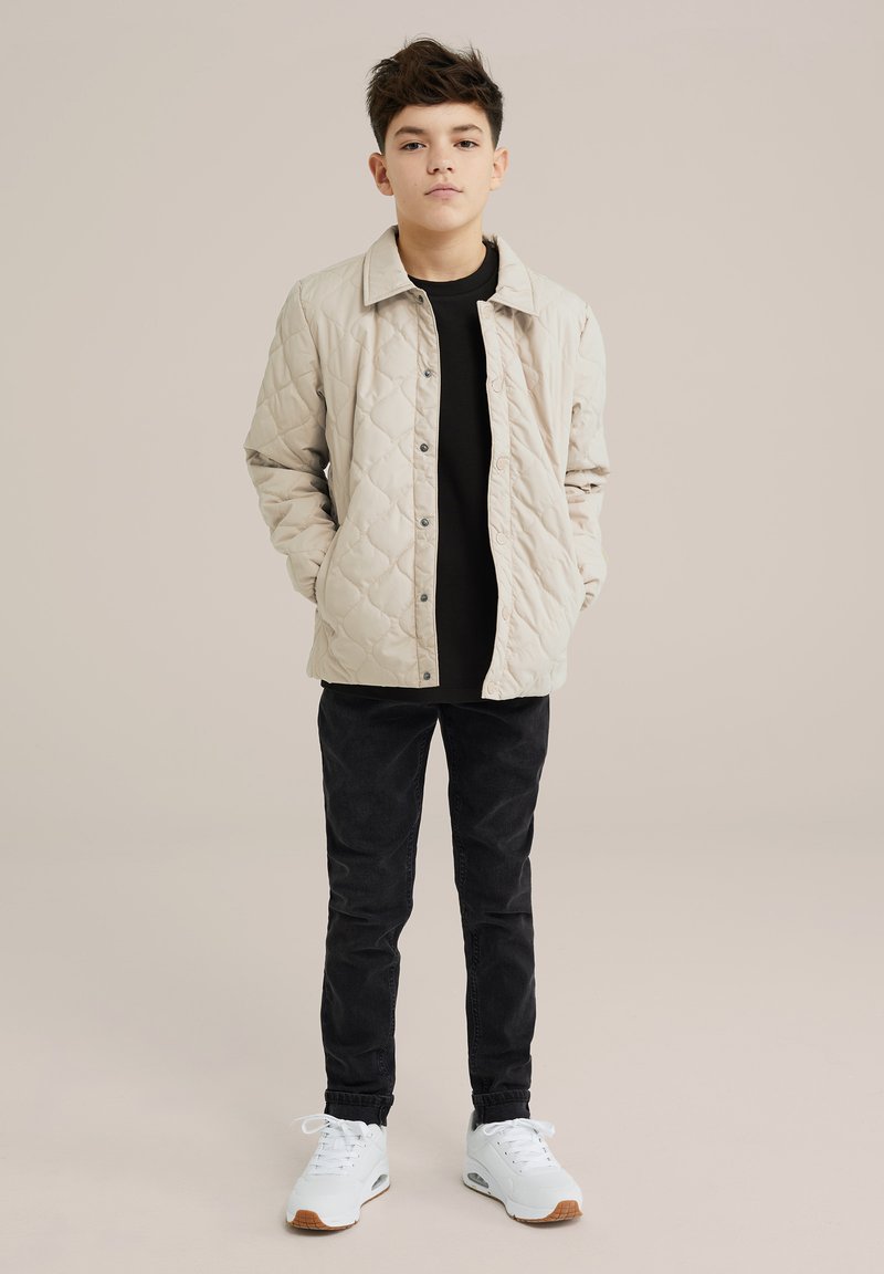 Beige pumpattu takki, jossa on nepparit, päällä musta paita. Mustat slim-fit-housut ja valkoiset tossut ruskeilla yksityiskohdilla täydentävät asun.