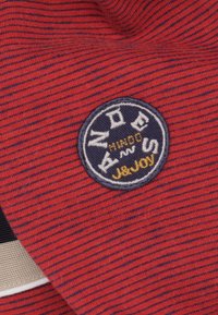 Rode en marineblauwe gestreepte stof met een ronde geborduurde logo die "AND" in het wit en "JOY" in het geel toont. Zachte textuur, casual design.