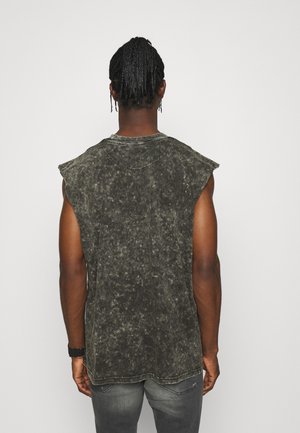 Uomo con capelli intrecciati che indossa una maglietta senza maniche nera e grigia scolorita e jeans grigi, in piedi davanti a uno sfondo bianco semplice.