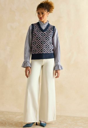 Navy patterned vest over striped shirt; wide, cream-colored pants; blue heels; textured fabrics; prominent v-neck and ruffled sleeves.
