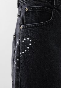Zwarte denim jeans met een zijzak versierd met een patroon van witte en zwarte rhinestones. Goudkleurige hardware zichtbaar op de zakken.