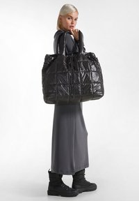 Borsa a spalla nera trapuntata con finitura in nylon lucido, doppi manici e chiusura con coulisse. Il modello indossa un vestito grigio e stivali neri.
