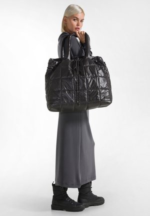Bolso tote negro acolchado con acabado de nylon brillante, asas dobles y cierre con cordón. La modelo lleva un vestido gris y botas negras.