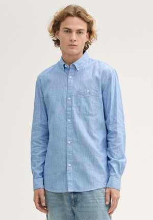 Light blue long-sleeve collared shirt made of textured fabric with a chest pocket and white buttons, paired with faded blue jeans.