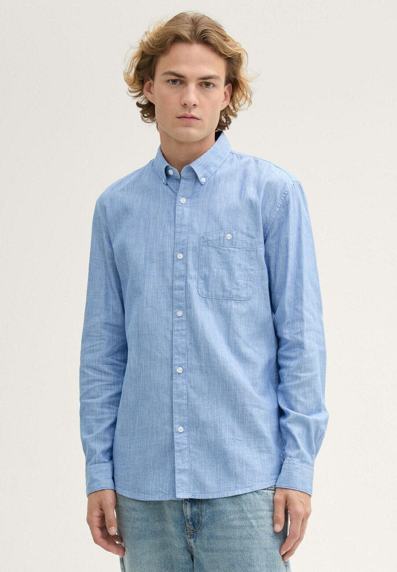 Helles blaues langärmliges Hemd mit Kragen aus strukturiertem Stoff, ausgestattet mit einer Brusttasche und weißen Knöpfen, kombiniert mit verwaschenen blauen Jeans.