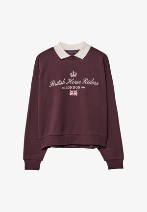 Bordo pulover s belim ovratnikom, ki na sebi nosi napis "Britanski konjeniški vozniki" in motiv krone. Vključuje tudi print Unije in napis "LONDON 1994".